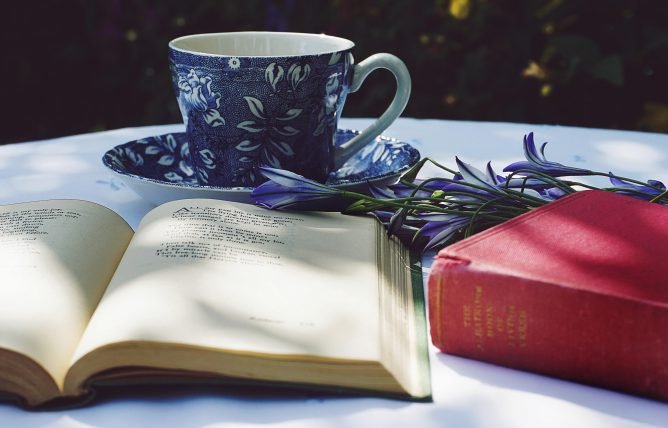 Book Opened on Top of White Table Beside Closed Red Book and Round Blue Foliage Ceramic Cup on Top of Saucer for World Poetry Day.